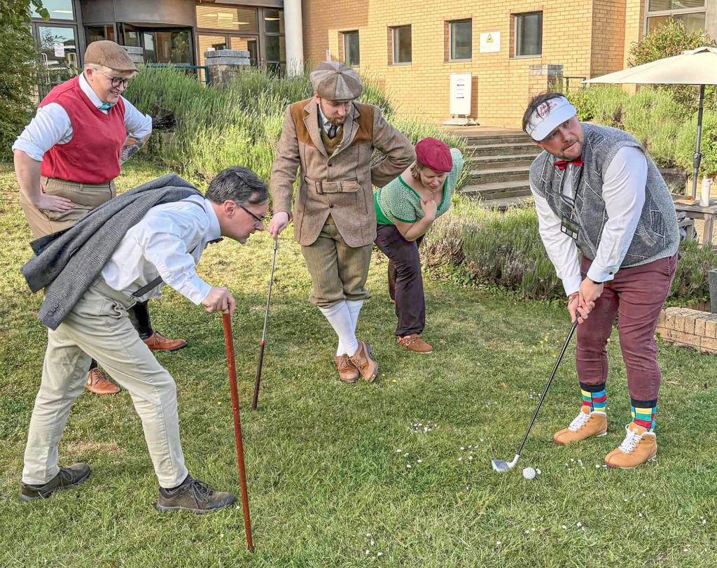 5 actors dressed like 1920s golfers pose on a golfing green